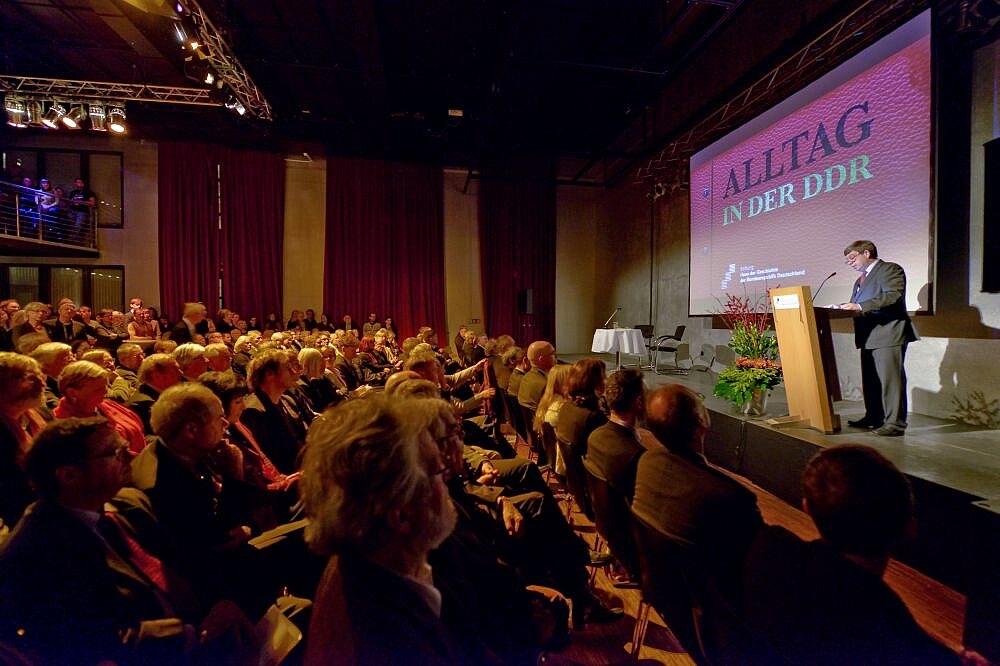 Feierliche Eröffnung des Museums in der Kulturbrauerei Stiftungspräsident Hütter auf einer Bühne, hinter ihm das Plakat der Ausstellung Alltag in der DDR, vor der Bühne sind Stuhlreihen mit Menschen