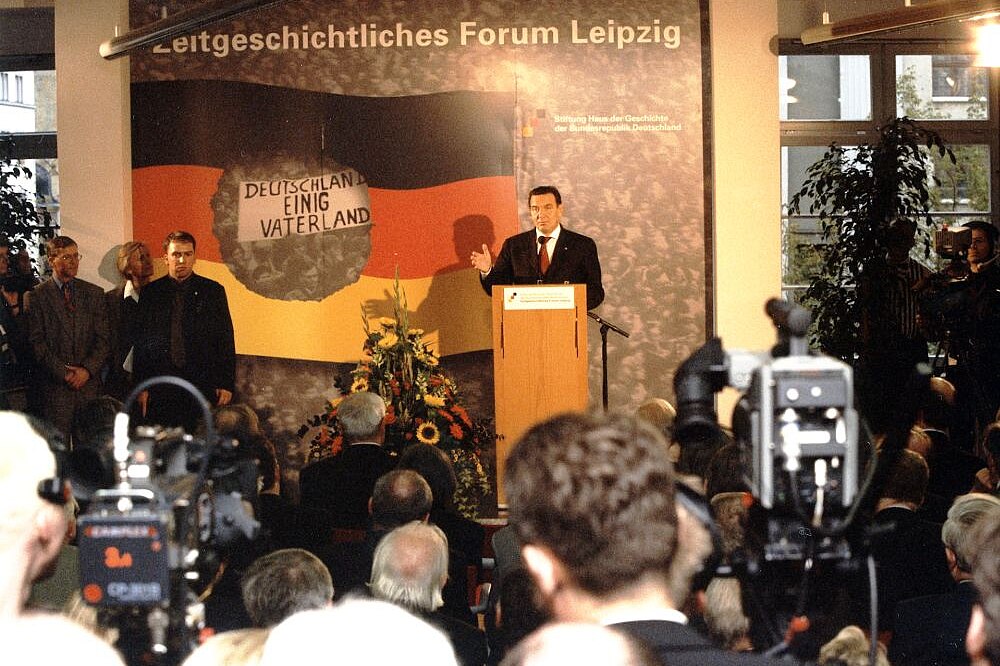 Der damalige Bundeskanzler Gerhard Schröder eröffnet das Zeitgeschichtliche Forum Leipzig Gerhard Schröder am Rednerpult, dahinter ein großes Plakat mit der Aufschrift Zeitgeschichtliches Forum Leipzig, vor ihm Besucher und Pressevertreter