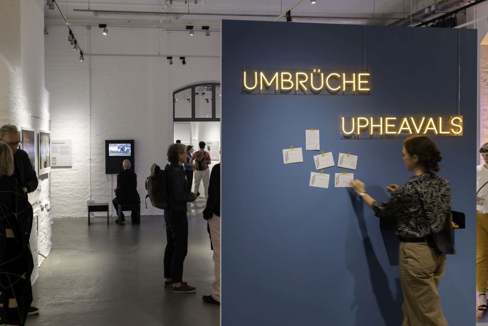 Besucherinnen und Besucher in der Ausstellung Besucherin vor einer Pinnwand mit dem Schriftzug "Umbrüche. Upheavals" in der Ausstellung.